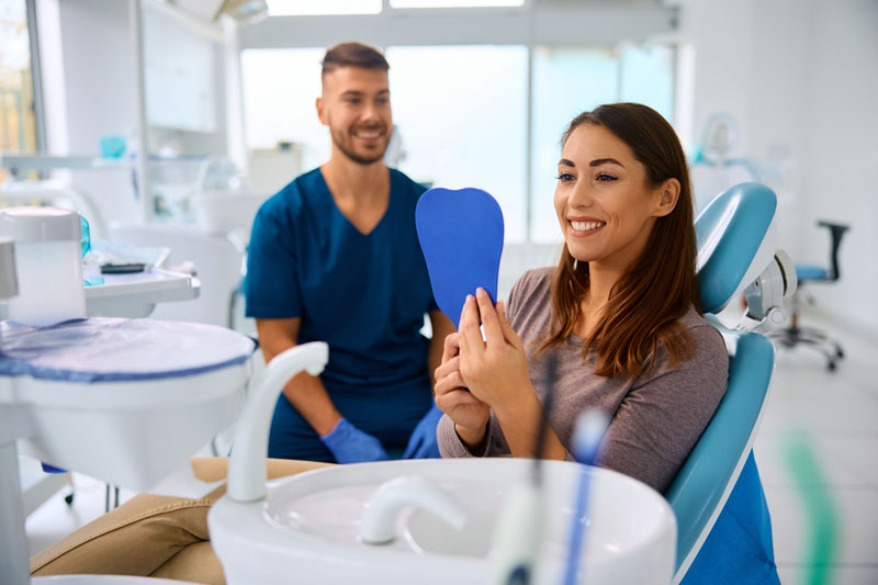 Happy woman looking at her teeth in a handheld mirror at dentist's office, male dentist interacting with the female patient